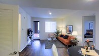 This single-floor space features a well-appointed open-concept living area that combines style and functionality. The living room is anchored by rich, dark hardwood-style flooring that extends throughout, creating a warm and cohesive aesthetic. Recessed ceiling lighting illuminates the space evenly, complemented by natural light streaming through a large window dressed with vertical blinds, which brightens the interior considerably.

The living area is generously sized, offering ample room for comfortable seating arrangements. A sliding glass door provides direct access to an outdoor area, enhancing the sense of openness and allowing for additional natural light. The clean white walls serve as a neutral backdrop, making the space feel airy and versatile.

Built-in media storage accommodates a wall-mounted television, while the open layout allows for flexible furniture placement. The space also includes a defined dining area, visible from the living room, reinforcing the open-plan design. Decorative touches such as indoor greenery and framed wall art contribute to the inviting, modern ambiance of this thoughtfully designed interior.