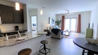 This single-floor unit is designed with an open-concept layout that seamlessly connects the kitchen, dining, and living areas, creating a spacious and airy feel throughout. The kitchen features granite countertops with a raised breakfast bar, pendant lighting overhead, and dark cabinetry that contrasts cleanly with the light-colored walls. Bar-style stools provide casual seating along the counter, making the space practical for everyday dining.

The living area is bright and welcoming, anchored by light wood-style flooring that extends throughout the main space. A ceiling fan with integrated lighting adds functionality, while large glass French doors dressed with coral-toned curtains invite natural light and lead to an outdoor area. The space is styled with neutral tones accented by navy blue, creating a cohesive and modern aesthetic.

A glimpse into the bedroom through an open doorway reveals a similarly bright and well-lit space. The overall ambiance of this unit is clean, contemporary, and functional, with thoughtful finishes and an open floor plan that maximizes the sense of space and connectivity between rooms.