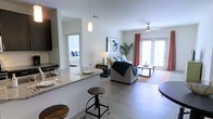 This single-floor unit is designed with an open-concept layout that seamlessly connects the kitchen, dining, and living areas, creating a spacious and airy feel throughout. The kitchen features granite countertops with a raised breakfast bar, pendant lighting overhead, and dark cabinetry that contrasts cleanly with the light-colored walls. Bar-style stools provide casual seating along the counter, making the space practical for everyday dining.

The living area is bright and welcoming, anchored by light wood-style flooring that extends throughout the main space. A ceiling fan with integrated lighting adds functionality, while large glass French doors dressed with coral-toned curtains invite natural light and lead to an outdoor area. The space is styled with neutral tones accented by navy blue, creating a cohesive and modern aesthetic.

A glimpse into the bedroom through an open doorway reveals a similarly bright and well-lit space. The overall ambiance of this unit is clean, contemporary, and functional, with thoughtful finishes and an open floor plan that maximizes the sense of space and connectivity between rooms.