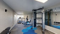 Located at 615 S Hardy Dr in Tempe, Arizona, this fitness center is a compact yet fully functional exercise room designed to support a comprehensive workout routine. The space features a distinctive flooring layout, combining standard carpet with bold, hexagonal blue tiles that add a modern, energetic aesthetic to the room. The walls are constructed of painted concrete block, lending a clean, durable feel, while a full-length mirror along one wall visually expands the space and assists with exercise form. Natural light filters in through windows and a glass-paneled door that opens to a view of lush greenery outdoors, keeping the atmosphere bright and inviting. Overhead, flush panel lighting ensures consistent illumination throughout the room. The fitness center is equipped with Matrix-branded cardio equipment, including multiple treadmills, an elliptical trainer, and a recumbent cycle. A Matrix functional trainer cable machine serves as the centerpiece for strength and resistance training, complemented by a weight bench for additional exercises. A ballet-style stretching bar and motivational wall art round out the space. The layout is efficiently organized to maximize usability, making this exercise room a well-appointed facility for a variety of fitness needs.