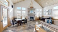 This 1110 square foot community room in Madison, WI, is a versatile and welcoming space designed for gatherings and events. The room boasts a vaulted ceiling with exposed wooden beams, creating an open and airy atmosphere. Large windows throughout flood the space with natural light and offer views of the surrounding area.

The room is anchored by a stone fireplace, adding warmth and a focal point to the space. Multiple seating areas are arranged throughout, including comfortable armchairs and round tables, perfect for small group discussions or casual dining. A large flat-screen TV mounted on the wall provides entertainment options.

The well-appointed kitchen features honey-colored wood cabinets, stainless steel appliances, and ample counter space. Tile flooring in the kitchen area transitions to a stylish wood-look flooring in the main room, unifying the space while defining different functional areas.

Decorative elements such as a model sailboat, coastal-themed artwork, and framed botanical prints add character and visual interest. The room's neutral color palette, primarily whites and warm woods, creates a calm and inviting environment suitable for various occasions.

Security and safety features are evident, with visible smoke detectors and emergency exit signs. The space is wheelchair accessible, with a door leading directly outside. Overall, this community room offers a blend of comfort, functionality, and aesthetic appeal, making it an ideal space for residents to gather, socialize, and host events.