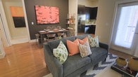 This well-designed single-floor space seamlessly integrates the living, dining, and kitchen areas into a cohesive open-plan layout, creating a sense of flow and connectivity throughout. The living area is anchored by a plush grey sofa adorned with a vibrant mix of coral, green, and neutral patterned throw pillows, complemented by a geometric area rug that adds texture underfoot. Warm hardwood flooring extends throughout the space, tying the zones together with a natural, inviting tone. A bold coral abstract artwork commands attention on the dark feature wall, serving as a striking focal point that bridges the living and dining areas. The dining zone accommodates a full-sized table with seating for multiple guests, ideal for entertaining. Transitioning into the kitchen, dark espresso cabinetry contrasts beautifully against stainless steel appliances, including a refrigerator and range, while mosaic tile backsplash details add a decorative finish. An open shelving unit provides both storage and display opportunities, enhancing the kitchen's contemporary aesthetic. The consistent use of dark tones, warm wood, and vivid accent colors throughout creates a modern, sophisticated ambiance with a welcoming energy.