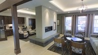 This thoughtfully designed single-floor space showcases a seamless open-concept layout that balances comfort and contemporary style. At its centerpiece stands a commanding double-sided fireplace clad in light-toned tile, rising to meet a tray ceiling adorned with recessed lighting and soft ambient LED strips that lend a warm, inviting glow to the entire area. The fireplace serves as a natural divider, elegantly separating a relaxed lounge area from a more formal dining space without sacrificing visual openness. The lounge area is furnished with plush armchairs and accented by rich dark wood doors, creating a cozy yet refined retreat. On the opposite side, the dining area features sleek wood-backed chairs and a round table, ideal for gathering. Natural light filters in through large windows dressed with floor-length drapes, adding warmth and a connection to the outdoors. The flooring transitions subtly between light hardwood-style surfaces and patterned area rugs, further defining each zone. Neutral wall tones in taupe and gray unify the space, while carefully placed artwork adds a curated touch. The overall ambiance is modern, polished, and welcoming — a well-appointed interior designed for both relaxation and social engagement.