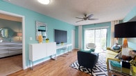 This single-floor unit is defined by its bold, consistent use of teal-toned walls that carry throughout every room, creating a cohesive and energetic atmosphere. The living area features wood-style flooring, a ceiling fan, and a sliding glass door that opens to a balcony, allowing natural light to fill the space. A built-in desk area along one wall offers a functional workspace, while the open layout provides ample room for seating arrangements.

The kitchen is well-appointed with dark cabinetry contrasted by white upper cabinets and marble-style countertops. Black appliances including a range and refrigerator are complemented by a large breakfast bar with seating for four, seamlessly connecting the kitchen to the living area. A stacked washer and dryer unit is also visible within the kitchen space.

The bedrooms continue the teal color scheme, with carpet flooring and ceiling fans providing comfort. One bedroom features bold red accent walls flanking the window, adding contrast and visual interest. Both bathrooms include dark wood vanities, marble-style countertops, and tub-shower combinations with decorative curtains and hardwood-style flooring.