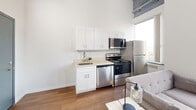 Located on the fourth floor of 601 Penn Street in Reading, PA, Unit 418 is a thoughtfully designed one-bedroom, one-bathroom residence that maximizes every square foot of its compact footprint. The open-concept living and kitchen area sets the tone with soaring ceilings that lend a sense of airiness well beyond the unit's physical size. Exposed industrial ductwork above adds character, nodding to the building's architectural heritage.

The kitchen is efficiently laid out along one wall, featuring white shaker cabinetry, light countertops, a stainless steel sink, and a full suite of stainless steel appliances including a refrigerator, electric range, microwave, and dishwasher. Large windows flood the living area with natural light and offer views of the surrounding cityscape.

The bedroom maintains the clean, minimal aesthetic with crisp white walls, warm hardwood-style flooring, and elevated ceilings. Recessed circular lighting adds a modern touch, while a baseboard heater ensures comfort year-round.

The bathroom is neatly appointed with a white tub/shower combination, a clean vanity with an integrated sink, a large mirror, and light tile flooring, all finished in a neutral gray and white palette. Hardwood-style flooring flows consistently throughout the main living spaces, tying the unit together with a warm, cohesive feel.