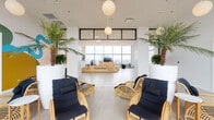 This elevated amenity space offers a compelling blend of modern design and resort-inspired comfort, situated high above the city with commanding panoramic skyline views through expansive floor-to-ceiling windows. The open layout is anchored by large-format light grey tile flooring that flows seamlessly throughout, creating a clean and cohesive aesthetic.

The main lounge area features symmetrically arranged rattan chairs with deep navy cushions, lending a relaxed yet refined atmosphere. Towering tropical palm trees in oversized white planters frame the space and reinforce its resort-like character. Spherical globe pendant lights suspended from the ceiling provide warm, ambient illumination throughout.

A standout architectural feature is the stepped, light wood platform installation that doubles as tiered seating and a communal gathering area. Black-trimmed details accent the natural wood tones, while striped cushions add a playful touch. An integrated flat-screen display and adjacent built-in planters make this area both functional and visually engaging.

A bold, colorful mural adds an artistic focal point, while black-framed glass partitions and doors elegantly define separate zones without sacrificing openness. One section of the space opens directly to an outdoor terrace with a pool, further extending the resort ambiance. The result is a thoughtfully designed, multi-functional space that balances style, comfort, and striking urban views.