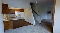 This two-story space offers a straightforward, functional layout across its main and upper levels. The main floor combines a kitchen and open living area in a practical arrangement. The kitchen is equipped with white appliances, including a refrigerator, electric range, and built-in microwave, all framed by warm wooden cabinetry. A peninsula with a light-colored countertop provides additional workspace and separates the kitchen from the adjacent living area. The kitchen features hardwood-style flooring, which transitions to plush carpeting in the living space. The living area is open and unfurnished, offering a neutral palette with light gray walls and carpet throughout. A staircase with carpeted treads and a painted white surround rises prominently along one wall, leading to the upper level. A single pendant light fixture hangs from the ceiling, providing a focal point overhead. The overall design is clean and understated, prioritizing practicality and ease of use. The neutral tones and simple finishes create a versatile backdrop suitable for a variety of furnishing styles.