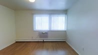 This single-floor space offers a clean, straightforward layout designed for comfortable everyday living. The main living area is characterized by warm hardwood-style flooring that runs throughout, lending a sense of continuity and warmth to the interior. A large double window dressed with vertical blinds serves as the focal point of the room, allowing an abundance of natural light to fill the space while providing adjustable privacy. The neutral, light-toned walls and white ceiling create a bright, open ambiance that makes the room feel airy and welcoming. Climate control is well-addressed, with a wall-mounted window air conditioning unit positioned beneath the windows for cooling, complemented by a baseboard heating system that runs along the perimeter of the room for warmth during cooler months. The clean lines and minimal detailing throughout give the space a practical, move-in-ready character. Overall, the layout is versatile and can easily accommodate a variety of furnishing arrangements, making it a functional and adaptable living environment.