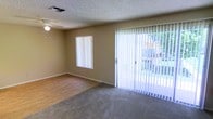 This single-floor residence offers a straightforward and functional layout designed for comfortable everyday living. The main living area is a notable feature of the space, combining two distinct flooring types — durable hardwood-style flooring in one section and plush carpeting in another — creating defined zones within an open setting. A ceiling fan is centrally mounted overhead, providing both lighting and air circulation throughout the room. The textured ceiling adds a classic touch to the interior. Natural light flows generously into the living area through a large sliding glass door fitted with vertical blinds, which also provides direct access to an outdoor area visible through the glass, where greenery and a fence can be seen. An additional window on the same wall further enhances the brightness of the room. The warm, neutral-toned walls throughout give the space a clean and inviting ambiance. HVAC vents are integrated into both the ceiling and lower wall areas, ensuring climate control across the living space. Overall, this residence presents a practical and well-lit environment with a welcoming atmosphere suited for a variety of living arrangements.
