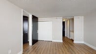 This single-floor unit offers a well-organized layout designed for functional, everyday living. Throughout the space, rich hardwood-style flooring provides a warm and cohesive foundation, complemented by neutral-toned walls and textured ceilings that add character to each room.

The kitchen is efficiently appointed with crisp white cabinetry extending from floor to ceiling, offering generous storage. Granite-look countertops surround a stainless steel sink, and a full-size refrigerator is neatly integrated into the compact layout, making the most of the available space.

The main living area is a standout feature, bathed in natural light from a wide bank of full-height windows fitted with horizontal blinds. These windows frame open views of the surrounding cityscape, lending an airy, elevated feel to the room. A baseboard heating unit runs along the wall, ensuring year-round comfort.

An entry corridor connects the living space to additional rooms, including what appears to be a closeted sleeping area and a bathroom featuring floor-to-ceiling tile work and a walk-in shower with mosaic tile flooring and grab bar fixtures. A large double-door closet in the entry zone provides ample storage. The overall ambiance is clean, understated, and move-in ready, with updated finishes throughout.