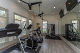 Located at 8064 S Fulton Pkwy in Fairburn, Georgia, this fitness center is a well-equipped, single-floor facility designed to support a comprehensive workout experience. The space features light wood-look flooring throughout, crisp white walls accented with crown molding, and recessed lighting complemented by ceiling fans that keep the environment comfortable during exercise.

Natural light floods the room through multiple large windows fitted with white plantation-style blinds, offering views of the surrounding greenery and creating an open, inviting atmosphere. A full-length wall mirror visually expands the space and allows users to monitor their form during workouts.

The facility is outfitted with a variety of cardio equipment, including Spirit-brand treadmills and an elliptical trainer, all positioned to take advantage of the natural light from the windows. A multi-station cable and weight machine provides strength training options, while a dedicated dumbbell rack offers free weight selections. Wall-mounted flat-screen televisions keep users entertained during longer sessions.

Decorative wall art in teal and white adds a touch of color and personality to the space. A dark-painted entry door with clearly marked exit signage and a fire safety panel ensure the facility meets safety standards. The overall ambiance is clean, bright, and functional, making it a welcoming environment for residents seeking a convenient on-site fitness option.