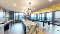 This single-floor space is a striking blend of luxury design and high-end entertainment functionality. The open layout flows seamlessly between a fully equipped kitchen, dining area, lounge, and game room, all unified by wide-plank hardwood flooring and a refined navy and gold color palette.

The kitchen is anchored by an oversized waterfall island clad in dramatic veined marble, providing generous counter seating along its length. Dark cabinetry lines the perimeter walls, complemented by a tiled backsplash, undermount sink, and integrated appliances. Sculpted gold-and-crystal chandeliers hang above, adding a bold artistic statement to the culinary space.

Adjacent to the kitchen, a dining area transitions naturally into the lounge and billiards zone. The game room is dominated by a sleek pool table positioned beneath a breathtaking canopy of cascading gold pendants that covers nearly the entire ceiling, creating an immersive, glamorous atmosphere. A plush sectional sofa faces floor-to-ceiling glazed doors that open to an outdoor balcony, framing panoramic city skyline views. The overall ambiance is one of refined opulence, designed for sophisticated entertaining at an elevated scale.