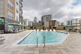Located at 515 Royal Court in the heart of Uptown Charlotte, NC, this outdoor amenity space offers a resort-style pool experience within an urban setting. The centerpiece is a generously sized rectangular swimming pool with crystal-clear blue water, finished with a clean, modern tile surround and stainless steel entry railings. The pool deck is paved with large-format stone tiles, providing a polished and durable surface throughout the space.

Surrounding the pool is a well-appointed lounge area featuring multiple chaise loungers and in-pool tanning ledge chairs, ideal for relaxing in the sun. Multiple seating clusters are shaded by vibrant green-and-white striped umbrellas, offering comfortable outdoor gathering spots. Lush greenery and manicured planters punctuate the perimeter, softening the urban environment and adding a welcoming atmosphere.

The pool is framed by the multi-story residential building, whose dark brick, warm wood-tone panels, and modern balconies create an intimate courtyard feel. On the opposite side, unobstructed views of the Uptown Charlotte skyline rise dramatically above the surrounding fence line, offering a striking urban backdrop. The overall ambiance blends contemporary design with practical outdoor living, making this pool deck a standout amenity in one of Charlotte's most vibrant neighborhoods.