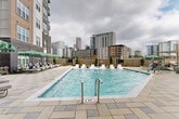 Located at 515 Royal Court in the heart of Uptown Charlotte, NC, this outdoor amenity space offers a resort-style pool experience within an urban setting. The centerpiece is a generously sized rectangular swimming pool with crystal-clear blue water, finished with a clean, modern tile surround and stainless steel entry railings. The pool deck is paved with large-format stone tiles, providing a polished and durable surface throughout the space.

Surrounding the pool is a well-appointed lounge area featuring multiple chaise loungers and in-pool tanning ledge chairs, ideal for relaxing in the sun. Multiple seating clusters are shaded by vibrant green-and-white striped umbrellas, offering comfortable outdoor gathering spots. Lush greenery and manicured planters punctuate the perimeter, softening the urban environment and adding a welcoming atmosphere.

The pool is framed by the multi-story residential building, whose dark brick, warm wood-tone panels, and modern balconies create an intimate courtyard feel. On the opposite side, unobstructed views of the Uptown Charlotte skyline rise dramatically above the surrounding fence line, offering a striking urban backdrop. The overall ambiance blends contemporary design with practical outdoor living, making this pool deck a standout amenity in one of Charlotte's most vibrant neighborhoods.