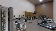 This fitness center offers a thoughtfully organized workout space designed to support a wide variety of exercise routines. The room features impressively high vaulted ceilings that lend an open, airy feel, complemented by neutral beige and warm tan walls that create a focused yet comfortable environment. The flooring is finished with durable carpet, providing a practical surface suited for gym use.

The strength training area is well-stocked with multiple weight benches, free weights, and a variety of resistance machines, catering to both beginner and experienced gym users. A large wall-mounted mirror spans a significant portion of the room, allowing users to monitor their form during exercises and visually expanding the sense of space. On the cardio side, the facility includes treadmills and additional aerobic equipment, ensuring a balanced fitness experience.

A wall-mounted television is positioned for easy viewing throughout the space, offering entertainment during workouts. Overhead fluorescent lighting ensures the entire room is evenly and brightly illuminated. The layout is spacious and thoughtfully arranged, allowing for comfortable movement between equipment. Overall, this fitness center provides a clean, functional, and fully equipped environment suitable for a comprehensive daily workout.