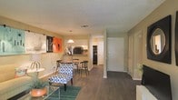 This single-floor residence offers a thoughtfully designed open-concept layout that seamlessly connects the living, dining, and kitchen areas into one cohesive living space. The living area features rich, dark hardwood-style flooring that anchors the room and contrasts beautifully with the warm, neutral-toned walls. A textured ceiling adds subtle depth overhead, while the open flow of the floor plan creates a sense of spaciousness and ease of movement throughout. The dining area is defined by a stylish pendant light that hangs above a compact dining table with coordinating chairs, making it an ideal setting for casual meals. Adjacent to the dining space, the kitchen is equipped with dark appliances including a refrigerator and oven, and features a convenient pass-through counter that maintains the open sightlines between the kitchen and main living area. Cabinetry in white provides a clean, bright contrast to the darker appliances. A front entry door is clearly visible, offering a well-defined entryway into the home. Colorful wall art adds personality and a pop of vibrancy to the living space. Overall, the residence combines functional design with a welcoming aesthetic, making it a comfortable and practical living environment.