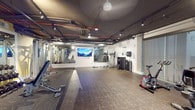 Located at 550 W Date St in San Diego's Little Italy neighborhood, this fitness center offers a well-equipped, single-floor workout facility designed to meet a wide range of exercise needs. The space features an industrial-inspired aesthetic, with an exposed ceiling showcasing ductwork and track lighting set against a dark finish, creating a modern and energetic atmosphere.

The cardio zone is outfitted with multiple treadmills and stationary bikes, including both upright and recumbent models, positioned along a glass-paneled wall that allows natural light to filter in from an adjacent corridor. This transparent boundary adds a sense of openness to the space.

The strength training area includes a comprehensive rack of dumbbells, an adjustable workout bench, and a cable/functional training machine, supported by wall-mounted mirrors that enhance the sense of depth and allow users to monitor their form. A large flat-screen TV is mounted on the wall, providing visual entertainment during workouts.

The open floor area, finished with durable rubber and patterned flooring, provides ample space for stretching, mat exercises, and functional movements. A ceiling fan aids in air circulation throughout the facility. The gym also offers easy access to an adjacent common area, rounding out a complete and convenient fitness experience.