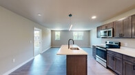This two-floor space showcases a thoughtfully designed modern interior with a cohesive aesthetic carried throughout. The main level centers around an open-concept kitchen and living area, where light wood-style flooring, recessed lighting, and large windows create a bright and airy atmosphere. The kitchen is well-appointed with dark wood shaker-style cabinetry, white quartz countertops, and a full suite of stainless steel appliances including a range, microwave, and refrigerator. A substantial center island with an undermount sink and matte black faucet serves as the focal point, complemented by a sleek linear pendant light overhead. A door from the main living area provides access to an outdoor balcony, adding connectivity to the exterior.

The upper level features carpeted bedrooms, one of which includes exposed ductwork lending an industrial touch, along with sliding barn door access to a walk-in closet. Bathrooms on both levels are finished with white quartz vanities, dark wood cabinetry, matte black fixtures, and backlit LED mirrors. One bathroom features a full tub-shower combination with large-format gray tile surround. Overall, the space balances contemporary and functional design elements throughout.