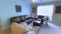 This single-floor space features a comfortable living area finished with soft wall-to-wall carpeting and painted in a calming light blue tone that contributes to an inviting, relaxed atmosphere. The room is furnished with a rich brown leather sofa set, including a matching armchair and ottoman, complemented by a cream-colored sofa in the foreground, offering ample seating throughout the space. A decorative floral wall plaque adds an artistic touch to the blue accent wall, while a framed artwork near the window provides additional character. Natural light filters generously into the room through a large window fitted with vertical blinds, brightening the interior. A small round side table positioned between the seating pieces provides functional surface space. Overhead, a ceiling-mounted light fixture with a classic multi-bulb design serves as the primary light source. A potted plant in the corner introduces a natural element, enhancing the overall warmth of the room. Crown molding along the ceiling adds a subtle architectural detail that elevates the finished look of the space.