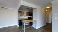 This residence at 800 City Tower Apartments offers a thoughtfully designed single-floor layout that balances functionality with modern style. The kitchen is a standout feature, framed by an elegant crown-molded archway that creates a clear yet open connection to the living area. Rich warm-toned cabinetry provides ample storage, while granite countertops lend a polished, upscale feel to the space. Black appliances, including a built-in microwave and range, complement the kitchen's cohesive aesthetic. Track lighting overhead ensures the kitchen is well-lit and practical. A movable granite-topped island on casters adds flexible prep and dining space, doubling as a functional centerpiece between the kitchen and living area. The flooring throughout features durable hardwood-style planks in a deep, warm tone that grounds the space with a sense of sophistication. Crisp white walls and white baseboards create a clean, bright contrast against the darker cabinetry and flooring, contributing to an overall ambiance that feels both inviting and contemporary. The thoughtful design details, from the crown molding to the seamless open-concept flow, make this a comfortable and stylish urban residence.
