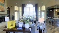 This single-story space makes a striking first impression with its soaring ceilings and dramatic arched window as the room's centerpiece. The oversized arch, flanked by tall, symmetrically placed windows dressed in navy blue patterned drapery, floods the interior with abundant natural light while offering views of lush outdoor greenery. Transom windows positioned above the doorways further enhance the bright, airy atmosphere throughout the space.

The interior is thoughtfully furnished with a refined, contemporary aesthetic. Seating arrangements include plush gray sofas and patterned accent chairs in coordinating blue and white tones, creating a cohesive and inviting palette. A dark wood coffee table anchors the central seating area, adorned with an elegant white ceramic vase holding fresh white calla lilies and yellow floral accents, adding a natural, decorative touch.

Layered table lamps provide warm ambient lighting, complementing the natural light. The overall ambiance balances openness with comfort, making the space feel both expansive and welcoming. The combination of tall architectural elements, curated furnishings, and abundant light gives this lounge a polished, hospitality-inspired character.