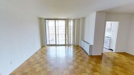 This single-floor space is characterized by its clean, minimalist design and an abundance of natural light. The main living area is finished with warm-toned parquet wood flooring laid in a classic diamond pattern, adding a timeless, elegant touch to the interior. Crisp white walls and smooth ceilings create a bright, airy atmosphere throughout.

A standout feature of the space is the expansive floor-to-ceiling sliding glass doors, which span nearly the entire width of the living area. These doors open onto a private balcony enclosed by a metal railing, offering open views of the surrounding urban landscape and allowing generous amounts of natural light to flood the interior. Sheer vertical drapes frame the glass doors, providing flexibility for light control and privacy.

A baseboard heating unit is positioned along the wall adjacent to the balcony doors, ensuring comfortable climate control. The open layout of the living area offers versatile arrangement possibilities, making it well-suited for a variety of uses. The overall ambiance is clean, neutral, and inviting, providing a functional and adaptable living environment.