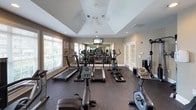 This fitness center is designed to support a comprehensive workout experience, offering a well-organized layout divided into distinct cardio and strength training zones. The main cardio area is generously sized, accommodating multiple treadmills, ellipticals, and stationary bikes arranged along a wall of large windows fitted with white horizontal blinds. These windows invite ample natural light and provide views of the surrounding greenery, creating an energizing atmosphere during workouts. A wall-mounted television is positioned for easy viewing from the cardio equipment.

The ceiling features a distinctive raised tray design with a ceiling fan at its center, enhancing the sense of vertical space and promoting air circulation throughout. Recessed lighting complements the natural light, ensuring the space is well-illuminated at all times.

The strength training area is equally well-appointed, featuring a cable machine, a dumbbell rack with a full range of weights, adjustable benches, and additional resistance machines. Full-length mirrors line the walls, visually expanding the space and allowing users to monitor their form. Durable rubber flooring covers the entire space, providing safety and comfort. A water dispenser and hand sanitizer station are conveniently available, adding a practical and hygienic touch to the facility.