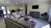 This single-floor space showcases a thoughtfully designed open-concept layout that seamlessly blends the living, dining, and kitchen areas into one cohesive environment. The living area is anchored by a plush gray sectional sofa and two white accent chairs arranged around a sleek, cylindrical metallic coffee table set atop a patterned area rug with warm red accents. A wall-mounted flat-screen television faces the seating arrangement, while a substantial gray multi-drawer dresser provides both storage and visual grounding to the space. A striking floral centerpiece adds a touch of natural elegance to the room. The kitchen, visible in the background, features white cabinetry, modern appliances, and under-cabinet lighting that enhances functionality while maintaining a clean aesthetic. Adjacent to the kitchen, a dining area with dark furnishings provides a defined space for meals. Recessed ceiling lighting throughout ensures the space is well-lit with a warm, inviting ambiance. The neutral white walls and smooth flooring create a bright, airy atmosphere, while the contemporary furniture choices and modern finishes reflect a refined, minimalist sensibility throughout.