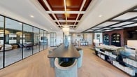 Located at 619 N Maple Ave in Ho-Ho-Kus, NJ, this thoughtfully designed club room and coworking space offers a sophisticated, multi-functional environment across a single expansive floor. The centerpiece of the space is a grand open area anchored by a striking coffered ceiling with rich mahogany wood inlays and an elegant brass globe pendant light, complemented by wide-plank herringbone hardwood flooring throughout. A long, dark conference table with curved upholstered chairs serves as the primary collaborative work surface, ideal for meetings or group work sessions. Adjacent to this is a welcoming lounge area featuring plush sectional sofas, a marble coffee table, and arched built-in cabinetry framing a fireplace and media wall, all bathed in natural light from large black-framed windows. A private meeting room enclosed by sleek steel-and-glass partition walls offers a quiet, gallery-like setting with a deep teal accent wall adorned with framed artwork. A well-appointed kitchen bar with a granite countertop, custom cabinetry, herringbone tile backsplash, and pendant lighting provides a refined refreshment area. Thoughtful seating nooks and transitional hallway spaces complete the layout, making this a versatile, design-forward destination for both focused work and social gathering.
