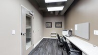This single-floor office space offers a functional and well-appointed environment designed for productive work. The room features warm taupe-toned walls paired with dark, multi-toned carpet tiles that add texture and a professional aesthetic underfoot. A built-in granite-topped workstation runs along one wall, providing ample surface area and accommodating a desktop computer, printer, and seating for multiple users via sleek black chairs. A compact white shelving unit offers additional storage and organizational flexibility. The ceiling showcases an industrial-modern aesthetic with exposed ductwork and structural beams finished in dark tones, complemented by recessed flat-panel LED lighting that ensures bright, even illumination throughout the space. Framed aerial stadium prints add a distinctive decorative touch to the walls. A glass-paneled interior door connects the office to an adjacent area, enhancing the sense of openness while maintaining privacy. White baseboards provide a clean contrast against the darker flooring, completing the polished look. Overall, the space balances a contemporary, professional atmosphere with practical design elements suited for daily office operations.