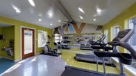 This fitness center offers a dynamic and motivating environment for residents to pursue their wellness goals. The space features a vaulted ceiling accented by exposed beams and a ceiling fan, creating an open, airy atmosphere enhanced by recessed lighting throughout. Bold, geometric wall art in yellow, taupe, and blue tones adds a lively visual energy to the room, while the vivid chartreuse and gray flooring reinforces the space's modern, spirited aesthetic. A full-length mirrored wall runs along one side of the room, reflecting the space and amplifying the sense of openness while allowing users to monitor their form during exercise. The fitness center is outfitted with a selection of cardio equipment, including treadmills and an elliptical machine, positioned to take advantage of both the mirrored wall and the natural light that filters in through the entry door. Motivational text is incorporated directly into the wall design, adding an inspiring touch to the overall environment. A wood-framed glass door provides access to the outdoors, contributing additional natural light and a connection to the exterior. The overall ambiance is energetic yet functional, making this fitness center a well-designed space for daily exercise routines.