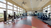 Located at 10327 W 52nd Pl in Arvada, Colorado, this well-appointed fitness center offers a comprehensive exercise environment designed to accommodate a variety of workout routines. The single-floor facility is characterized by its notably high ceilings, which lend an open, airy atmosphere throughout the space. Large, multi-pane windows line the walls, flooding the interior with abundant natural light and providing views of the surrounding outdoor landscape.

The flooring features a distinctive multi-colored rubber surface with sweeping geometric patterns in red, tan, and gray, providing both visual appeal and a durable, cushioned workout surface. Ceiling fans are distributed throughout to maintain comfortable airflow, complemented by linear overhead fluorescent lighting for consistent illumination.

The fitness center is thoroughly equipped with a broad selection of cardio machines, including treadmills, ellipticals, stationary bikes, and stair climbers, positioned to take advantage of the natural light from the windows. A dedicated strength training area includes cable machines, weight benches, free weights, kettlebells, and resistance equipment. Stability balls and additional functional fitness accessories are neatly organized along the walls. Wall-mounted televisions and full-length mirrors further enhance the workout experience, while posted fitness guidelines and instructional charts provide useful reference for users.