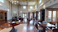 Located at 5701 Saratoga Blvd in Corpus Christi, Texas, this single-floor community facility is defined by its dramatic double-height ceilings and an abundance of natural light. The grand main lounge serves as the heart of the space, showcasing rich hardwood flooring, a coffered ceiling with recessed lighting, and clerestory windows that flood the interior with daylight. A modern chandelier anchors the room, while a built-in fireplace with bold abstract artwork above it adds a refined focal point. Comfortable seating arrangements throughout encourage gathering and relaxation. Floor-to-ceiling drapes frame expansive glass doors and windows that open to views of the outdoor pool area, seamlessly connecting interior and exterior spaces. A dedicated seating nook near the pool-facing windows offers an intimate retreat with ample natural light. The kitchen is well-appointed with rich cherry wood cabinetry, granite countertops, a tile backsplash, and full appliances, providing full functionality for events or daily use. Additional areas include a workstation or leasing office zone with a wall-mounted television, open shelving, and adjacent private office spaces. The overall ambiance is polished yet welcoming, combining contemporary design with warm, residential-style finishes throughout.