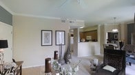 This single-story space showcases an open-concept design that seamlessly connects the living area to the kitchen, creating a sense of spaciousness and flow. The living area features soaring vaulted ceilings that enhance the airy ambiance, complemented by neutral warm beige walls that provide a versatile backdrop for furnishings and décor. Light-toned carpeting anchors the living space, transitioning to tile flooring near the kitchen entry.

The living area is tastefully appointed with decorative elements, including framed botanical artwork, tall sculptural vases, and floral arrangements that add a refined touch. A ceiling fan overhead provides both form and function.

The kitchen is positioned at the rear of the open floor plan, partially visible through a half-wall peninsula that defines the space while maintaining visual connectivity. It features wood-toned cabinetry, a white refrigerator, and a microwave, with overhead lighting illuminating the workspace. A pass-through window allows natural light to filter into the kitchen from an adjacent window.

Crown molding details throughout add an element of architectural sophistication, while the cohesive neutral palette creates a calm and welcoming overall ambiance.
