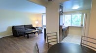 This single-floor space offers a practical and welcoming layout with a seamless flow between the living area and kitchen. The living room is furnished with a dark sofa, a coffee table, and a cushioned armchair, all set atop warm hardwood-style flooring. A floor lamp and a bright window with light curtains provide a well-lit, comfortable atmosphere. A structural column subtly divides the living space from the kitchen area, adding architectural interest while maintaining an open feel. The kitchen features dark cabinetry, a countertop with a sink, and a window overlooking lush greenery, filling the space with abundant natural light. The transition from hardwood-style flooring in the living area to tile in the kitchen clearly delineates the two functional zones. A dining area with a round table and metal-framed chairs is conveniently positioned between the kitchen and living room, making it ideal for casual meals. The overall ambiance is clean, neutral, and functional, with light-toned walls complementing the darker furnishings and cabinetry throughout. Ceiling fans contribute to the comfort of the space, making it both practical and inviting.