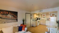This single-floor residence offers a well-organized and efficiently designed layout that makes thoughtful use of its available space. The open-concept living and kitchen area forms the heart of the home, where warm light wood flooring runs seamlessly throughout, contributing to a bright and cohesive atmosphere.

The living area is furnished with a comfortable white sofa accented by colorful throw pillows in orange and blue, adding a lively touch to the space. Bold framed artwork on the walls, including a striking train station print, gives the room character and a sense of urban style.

The kitchen is positioned in an open alcove adjacent to the living area, featuring light wood cabinetry, a white electric range, a microwave, and ample counter space. The layout is practical and accessible, with under-counter storage and a clean, neutral finish.

A separate dining nook accommodates a compact white table with matching white slatted chairs, creating a defined space for meals. Framed artwork and a small decorative shelf with a floral arrangement lend warmth to this corner. A television and media console are positioned nearby, adding to the functionality of the dining and entertainment area.

A hallway visible from the main living space leads to additional areas, including what appears to be a bathroom, maintaining a clear and logical flow throughout the residence.