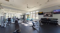 This single-floor fitness center is designed to support a comprehensive workout experience, combining thoughtful architectural details with a wide array of equipment. The space is finished with soft blue-gray walls, white crown molding, and dark rubber flooring that provides both durability and comfort underfoot. Ceiling fans are positioned throughout to promote air circulation, and recessed lighting supplements the abundant natural light that streams in through the large, multi-pane windows lining multiple walls, offering views of the surrounding outdoor landscape.

The cardio zone features treadmills, elliptical trainers, and a rowing machine, all positioned to take advantage of the expansive window views. Full-length mirrors are integrated along one wall, visually expanding the space and aiding form during exercise. The strength training area is well-appointed with a multi-station cable and selectorized machine system, a Smith machine, and various selectorized strength machines. A dedicated free weights section includes a tiered dumbbell rack with a full range of weights and adjustable benches. Stability balls and medicine balls add functional training options to the layout.

A TRX suspension training system is wall-mounted near the entrance, alongside a white storage credenza and posted fitness center rules. Wall-mounted flat-screen televisions provide entertainment throughout the space, contributing to an energizing and functional training environment.