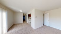 This single-floor residential unit offers a practical and well-organized layout designed for comfortable everyday living. The living area is generously sized, featuring neutral beige carpet, crisp white walls, and a large sliding glass door fitted with vertical blinds that floods the space with natural light and provides views of the surrounding exterior. A wall-mounted HVAC unit ensures climate control year-round.

The kitchen is thoughtfully configured with warm-toned wood cabinetry, light granite-style countertops, and white appliances including a gas range and a full-size refrigerator. Light tile flooring and a fluorescent overhead fixture keep the kitchen bright and easy to maintain. A closet with open shelving is conveniently located adjacent to the kitchen area.

The bedroom is a comfortable, carpeted space with a window that brings in natural light. A closet with double doors provides ample storage. An additional closet near the entry area offers even more organizational flexibility.

The bathroom is clean and functional, finished in crisp white tones with a shower/tub combination, toilet, and towel bars. The neutral palette throughout the unit creates a cohesive, move-in ready atmosphere that is easy to personalize.