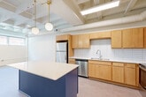 Located at 8 S Sixth St in Council Bluffs, Iowa, this 2-bedroom, 1-bathroom unit showcases an industrial-inspired design highlighted by exposed ceiling beams, ductwork, and structural elements painted crisp white throughout. The open kitchen is a standout feature, outfitted with warm maple cabinetry, a white tile backsplash, stainless steel appliances including a refrigerator, dishwasher, and range, and a bold navy blue island with a white countertop topped by globe pendant lighting — creating a stylish, functional focal point. The adjoining living area flows seamlessly from the kitchen, offering an open and airy feel. Both bedrooms are carpeted for comfort; the primary bedroom is a well-proportioned space featuring windows that draw in natural light, while the second bedroom is cozy and versatile. A dedicated closet provides built-in wire shelving for organized storage. The full bathroom features a wood-toned vanity with ample drawer storage, a large mirror with modern globe sconce lighting, and a full tub-shower combination. A connecting hallway ties all rooms together efficiently. The exposed-ceiling aesthetic lends the unit a loft-like character, making it a distinctive and functional residence.