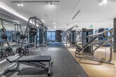 This sophisticated fitness facility offers a spacious, well-appointed environment designed to support a full range of workout activities across a single open floor. The space is divided into distinct functional zones, each thoughtfully designed with premium materials and modern aesthetics throughout.

The cardio zone lines the perimeter near expansive floor-to-ceiling windows, flooding the area with natural light and offering views of the surrounding cityscape. A row of treadmills and stationary bikes — equipped with individual digital screens — provides a high-end workout experience. Lush indoor greenery with ambient blue lighting adds a striking visual element, creating a fresh and energizing atmosphere.

The strength training area features a comprehensive selection of cable machines, free weights, and resistance equipment, set against a decorative botanical mural wall that adds an artistic touch to the space. Rubberized flooring in this zone provides both safety and durability.

A separate studio space is defined by sleek black-framed glass partitions with patterned detailing, offering a versatile area ideal for group classes or personal training. Full-length mirrored walls and linear ceiling lighting enhance the sense of openness. Light wood-tone flooring runs throughout the main zones, complementing the clean white walls and creating a cohesive, modern aesthetic. A water station and building management panel are also conveniently located within the facility.