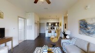 This single-floor residence offers a seamless open-concept layout that connects the living, dining, and kitchen areas into one cohesive and inviting space. The living area is anchored by rich dark hardwood-style flooring, a ceiling fan mounted to soaring vaulted ceilings, and an abundance of natural light that creates a warm, airy ambiance. A tasteful area rug and contemporary furnishings, including a distinctive egg-style accent chair and a comfortable sofa, complete the living space.

The kitchen is a standout feature, showcasing dark cabinetry paired with crisp white countertops and a breakfast bar with barstool seating — ideal for casual dining or entertaining. Pendant lighting and track lighting illuminate the kitchen area, adding both function and modern flair. Black appliances provide a sleek contrast to the light countertops and backsplash.

The overall design aesthetic is clean and contemporary, with neutral white walls serving as a versatile backdrop for the rich tones of the cabinetry, flooring, and accent decor. Thoughtful touches such as wall art and decorative lighting contribute to a polished, move-in-ready feel. The open flow and well-proportioned layout make this space as functional as it is visually appealing.