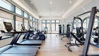 This fitness center is a thoughtfully designed exercise space that offers a full suite of equipment to support a wide range of workout routines. The room features a raised tray ceiling with recessed lighting and a large ceiling fan, contributing to an open, airy ambiance. Light hardwood-style flooring runs throughout, providing a clean and durable surface underfoot.

A continuous row of large, dark-framed windows lines the perimeter walls, flooding the space with natural light and offering views of the surrounding outdoor area, including a pool and neighboring buildings. The windows are fitted with roller shades, allowing for adjustable light control.

The cardio section is well-stocked with multiple treadmills, elliptical machines, a stair climber, stationary bikes, and a rowing machine. The strength training area features a cable machine, a multi-function rack with a bench press setup, adjustable benches, and a comprehensive dumbbell rack. Full-length mirrors are mounted along one wall, enhancing the sense of space while allowing users to monitor their form.

A built-in shelving unit and a small decorative nook are positioned along one interior wall, adding a polished finishing touch. The overall design is modern and functional, offering a well-rounded fitness environment with an inviting, light-filled atmosphere.