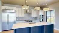 This well-appointed 2-bedroom, 3-bathroom single-story home offers a generous and thoughtfully designed floor plan. The standout kitchen features a large navy blue island with seating, contrasted by crisp white cabinetry, a subway tile backsplash, stainless steel appliances including a built-in refrigerator and double wall ovens, a gas cooktop, and a statement range hood. Three coordinating drum pendant lights illuminate the island, adding a refined touch to the space. Light hardwood flooring flows throughout the main living areas, creating a warm and cohesive aesthetic. Two entrance points provide convenient access to the home. The primary bedroom is a comfortable retreat with sliding glass doors opening to a private balcony with outdoor views, while the second bedroom features a ceiling fan and a large window for natural light. A stylish barn door with matte black hardware adds character to the hallway. The walk-in closet provides ample built-in shelving and hanging storage on multiple sides. The bathrooms are finished with large-format neutral tiles, glass-enclosed walk-in showers, and classic vanities with detailed cabinetry. Neutral gray walls and recessed lighting throughout contribute to the home's clean, contemporary ambiance.