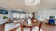 Located in Kissimmee, Florida, this single-floor space offers a well-organized layout that includes a living room, a combined dining room and kitchen area, and a secondary living space. Rich hardwood-style flooring runs throughout, providing a warm and cohesive look across all areas.

The main living room is generously sized, featuring vaulted ceilings, soft blue and grey wall tones, and multiple sets of white-framed French doors that invite natural light and provide outdoor access. A wall-mounted television and built-in ledge serve as a focal point, while comfortable seating arrangements create an inviting atmosphere. Large glass-paneled doors on both sides of the room offer views of the surrounding exterior.

The dining area is bright and open, anchored by a round wooden dining table beneath a ceiling fan. A dramatic arched window above the French doors floods the space with natural light, and vaulted ceilings add a sense of height and openness. Tall indoor plants enhance the airy ambiance.

The kitchen features white cabinetry, a full-size refrigerator, and a large wrap-around peninsula with a light-colored countertop, offering both workspace and a natural divider between the kitchen and the adjacent living areas. Recessed lighting and a window above the sink keep the kitchen bright and functional. French doors in the kitchen also provide direct outdoor access, reinforcing the seamless indoor-outdoor flow throughout the space.