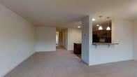 This well-appointed multi-story home spans three floors, offering a thoughtfully designed layout that balances open communal spaces with practical functionality. The main living area is spacious and inviting, featuring plush wall-to-wall carpeting in a warm neutral tone that adds comfort underfoot. Recessed lighting throughout the ceiling ensures consistent, even illumination, complementing the bright and airy atmosphere created by natural light filtering in through windows near the entryway.

The kitchen is positioned in an open-concept arrangement adjacent to the living area, separated by a structural column that adds architectural character. It features rich, dark wood cabinetry, a stone-topped breakfast bar with seating overhang, and pendant lighting that adds a refined touch above the counter. Black appliances anchor the space, providing a clean contrast to the lighter countertops and walls.

A hallway connects the main living areas to additional rooms, with a closet visible and a front entry door that reinforces the home's accessibility and flow. The clean white walls and neutral palette throughout create a versatile backdrop that suits a variety of interior styles. With three full floors, this home provides ample room for living, working, and entertaining across multiple levels.