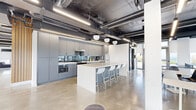 Located at 333 University Avenue in Kingston, Ontario, this single-floor commercial space offers a generous open-plan layout with a distinctly modern industrial aesthetic. Polished concrete floors, exposed ductwork, and dark ceilings with suspended linear LED lighting run throughout, creating a cohesive and contemporary atmosphere.

The fully equipped kitchen is a standout feature, outfitted with floor-to-ceiling grey cabinetry, a dark gloss tile backsplash, stainless steel appliances including a range, dishwasher, microwave, and full-size refrigerator, and a large central island with seating for multiple guests. Globe pendant lights add a refined touch overhead.

The open lounge and collaborative areas are defined by floor-to-ceiling windows along multiple walls, flooding the space with natural light and offering panoramic views of the Kingston cityscape and surrounding greenery. Flexible seating arrangements — including modular lounge furniture, round café tables, and bistro chairs — support a variety of uses. A wood-slatted accent wall introduces warmth against the industrial palette, while colorful modular carpet tiles delineate distinct activity zones. The space is well-suited for professional, hospitality, or co-working purposes.