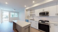 This single-floor space is designed with a clean, contemporary aesthetic throughout. The kitchen serves as the heart of the unit, showcasing white shaker-style cabinetry with matte black hardware, a classic white subway tile backsplash, and light stone countertops that extend across a generously sized center island — ideal for meal prep or casual dining. A full suite of stainless steel appliances includes a gas range, over-the-range microwave, dishwasher, and a French door refrigerator. Open shelving provides both functional storage and display space, while recessed lighting and light hardwood flooring create a bright, welcoming atmosphere. The kitchen flows naturally into the open living area, which continues the hardwood flooring and neutral white walls, lending the space a sense of openness and versatility. The bathroom is finished with large-format beige floor tiles and features a soaking tub enclosed within a tiled surround with glass sliding doors, complemented by a clean white vanity. A carpeted bedroom offers a slide-in closet with mirrored doors, providing comfortable and quiet retreat. Throughout the space, recessed lighting, a ceiling fan, and a consistent neutral palette contribute to a cohesive, modern living environment.