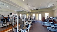 The fitness center at 825 E Palomar in Chula Vista is a well-appointed, single-floor facility designed to support a comprehensive workout experience. The space is outfitted with a variety of strength training machines arranged in an organized layout, providing ample room to move between stations comfortably. Large wall-to-wall mirrors line one side of the room, visually expanding the space and allowing users to monitor their form during exercise. Recessed lighting and multiple ceiling fans are installed throughout, ensuring the room remains well-lit and properly ventilated during workouts. Natural light enters generously through double French doors at the far end of the room, which also offer convenient access and contribute to an open, airy atmosphere. The flooring consists of durable, dark carpeting suited for fitness use. The overall aesthetic features neutral tones with clean crown molding details along the ceiling, lending the space a polished and well-maintained appearance. The combination of functional equipment, thoughtful layout, and bright, comfortable surroundings makes this fitness center a practical and inviting amenity.