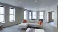 This single-floor space features a well-appointed living area designed with a clean, modern aesthetic. The room is finished with smooth grey walls and recessed ceiling lighting that creates an even, ambient glow throughout the space. Large, dark-framed windows line the walls in a slight angular arrangement, flooding the interior with natural light and framing views of the surrounding urban cityscape. The windows serve as a defining architectural feature, reinforcing the connection between the interior and the city beyond.

The living area is generously proportioned, providing ample room for comfortable furniture arrangement. A cream-toned L-shaped sectional sofa with warm orange accent pillows anchors the space, complementing the neutral color palette. A low white coffee table sits at the center, adding a functional yet minimalist element to the layout. Soft carpet flooring in a muted grey tone adds warmth underfoot and contributes to the overall understated, sophisticated atmosphere.

The overall ambiance is calm and refined, with a cohesive palette of cool greys and whites punctuated by warm accent tones. The space feels open, airy, and thoughtfully designed for both relaxation and everyday living.