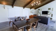 This single-floor space showcases a striking mid-century modern design, highlighted by beautiful exposed wood beam ceilings with warm-toned planking that lend the interior a rustic yet refined character. The open-plan layout seamlessly connects the dining and living areas, creating a cohesive and inviting atmosphere throughout.

The dining area is generously appointed with two large dark wood tables surrounded by metallic bronze industrial-style chairs, offering ample seating for gatherings. Overhead, a sculptural multi-globe chandelier serves as a standout focal point, adding a contemporary artistic touch to the space.

Adjacent to the dining area, the living space is comfortably arranged with a large sectional sofa in charcoal gray, accented by a bold orange sofa that introduces a vibrant pop of color. A wall-mounted flat-screen television and a decorative geometric wall art arrangement complete the living area, contributing to the space's modern sensibility.

Light-toned hardwood flooring runs throughout, complementing the natural wood ceiling and tying the design together harmoniously. Vertical blinds on the windows provide privacy while allowing for flexible light control. Overall, the space exudes a warm, casual, and well-curated ambiance ideal for both relaxing and entertaining.
